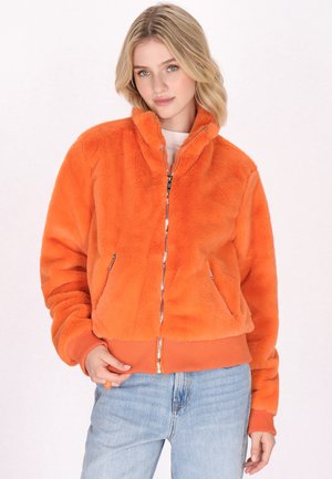 Veste en fausse fourrure orange avec fermeture éclair, poches latérales et poignets et ourlet côtelés. Coupe courte et décontractée. Portée sur un haut léger et un jean bleu.
