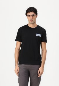 Sort bomulds T-shirt med rund halsudskæring og korte ærmer. Har et lille blåt og hvidt patch på venstre brystområde.