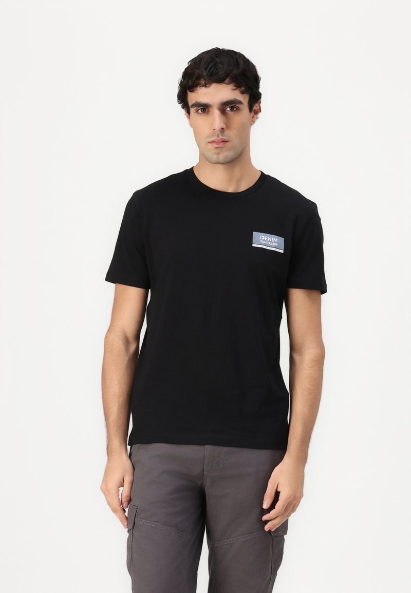 Sort bomulds T-shirt med rund halsudskæring og korte ærmer. Har et lille blåt og hvidt patch på venstre brystområde.