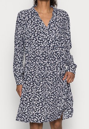 Mujer con un vestido de manga larga azul marino con un pequeño patrón floral blanco y un cuello en V, de pie frente a un fondo claro.