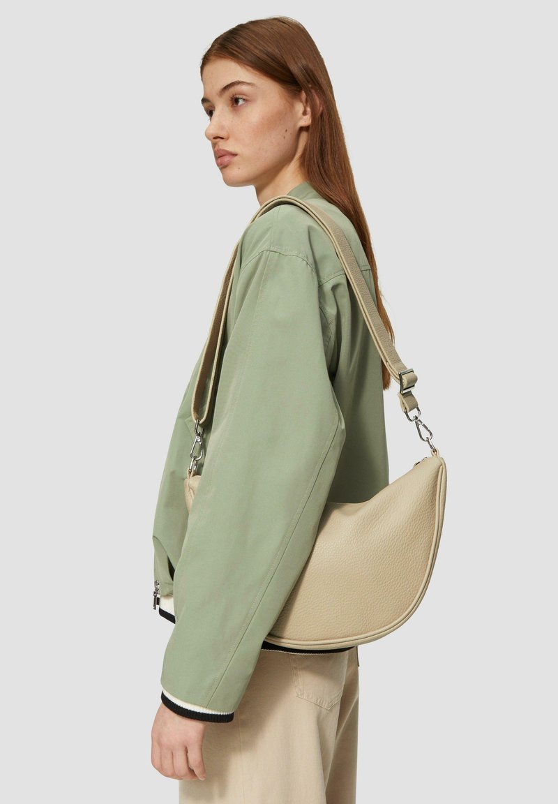 Sac à bandoulière en cuir beige avec une forme incurvée, finition texturée, sangle réglable et quincaillerie en argent, porté de manière décontractée sur une veste verte.