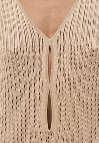 Κοντινή λήψη μπεζ πλεκτού υφάσματος με κάθετο ριμπ μοτίβο και δύο οβάλ ανοίγματα δημιουργώντας σχέδιο κλειδαρότρυπας στην περιοχή του στήθους.