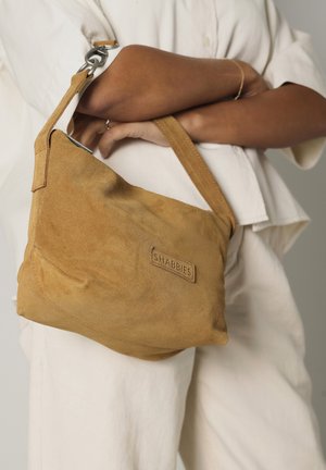 Tan suede shoulder bag with a textured surface, adjustable strap, and a logo patch. Curved shape and prominent stitching details.