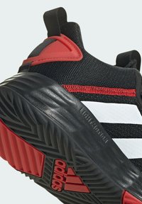 Zapatilla deportiva negra con acentos rojos, con una parte superior texturizada, tres franjas blancas y una suela negra con patrón que presenta la marca "adidas".
