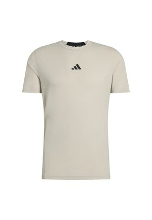 Jasnobeżowa koszulka z krótkim rękawem wykonana z bawełny, z czarnym logo Adidas na piersi. Okrągły dekolt i standardowy krój.