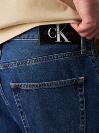 Mörkblå denimjeans med en svart läderlapp som har vit text "CK" och "Calvin Klein Jeans" på midjebandet.