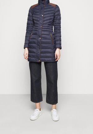 Marineblauer Puffer-Mantel mit hohem Kragen, braunen Schulterakzenten und mit Reißverschluss versehenen Taschen; kombiniert mit weit geschnittenen dunklen Jeans und weißen Sneakers.
