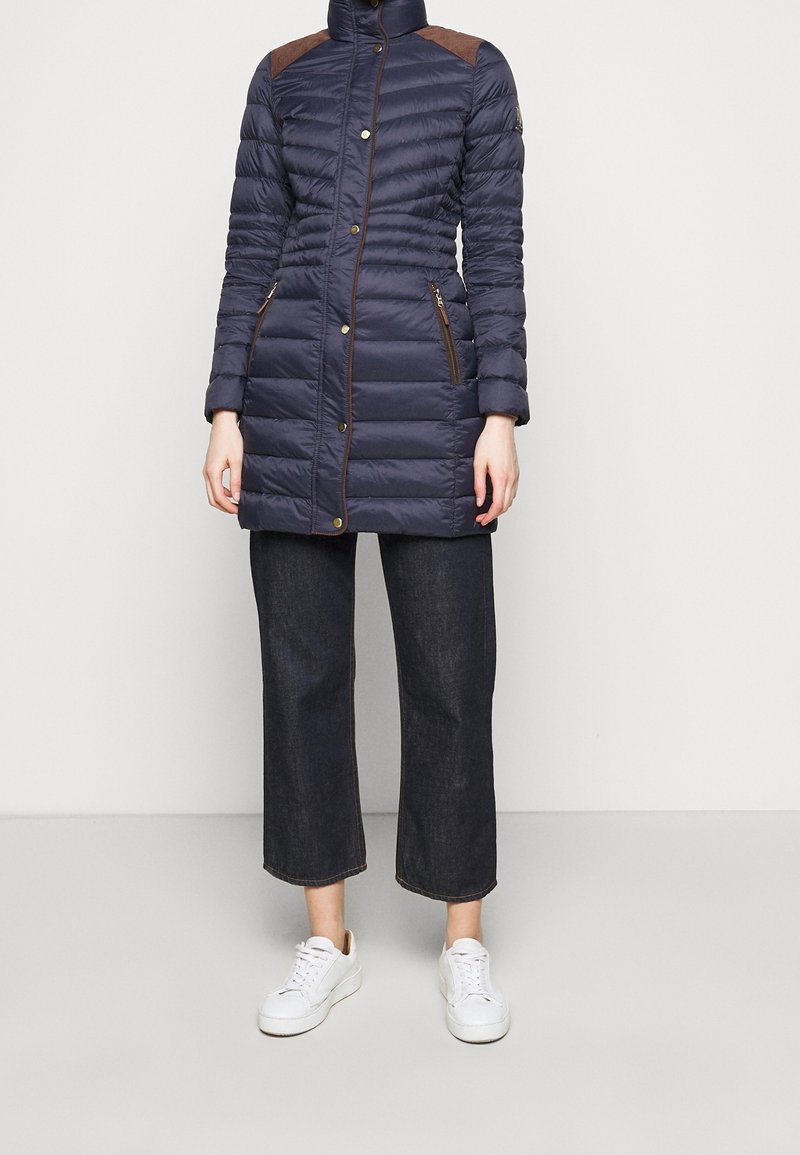 Mörkblå puffjacka med hög krage, bruna axelaccenter och fickor med dragkedja; tillsammans med vida mörkdenim och vita sneakers.