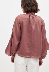 Vätskefylld, rosafärgad blus med vida ärmar, ett nyckelhål bak, och samlade detaljer vid ärmsluten, bärs över vita byxor.