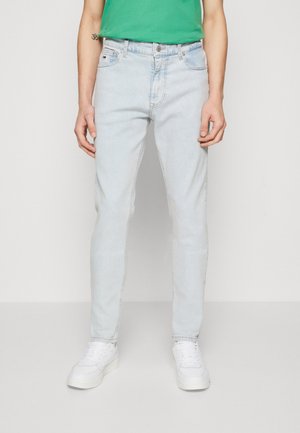 Man draagt lichtblauwe slim-fit jeans, witte sneakers en een groen overhemd, staand tegen een effen lichte achtergrond.