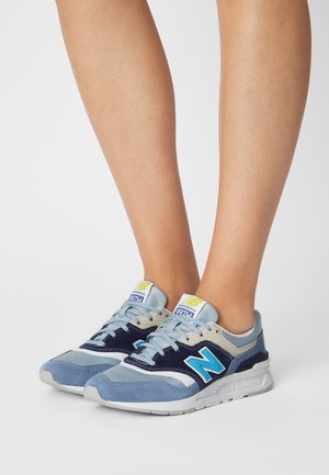 Jambes portant des baskets New Balance bleues et grises avec des lacets bleu clair et un logo "N" bleu vif sur des semelles blanches.