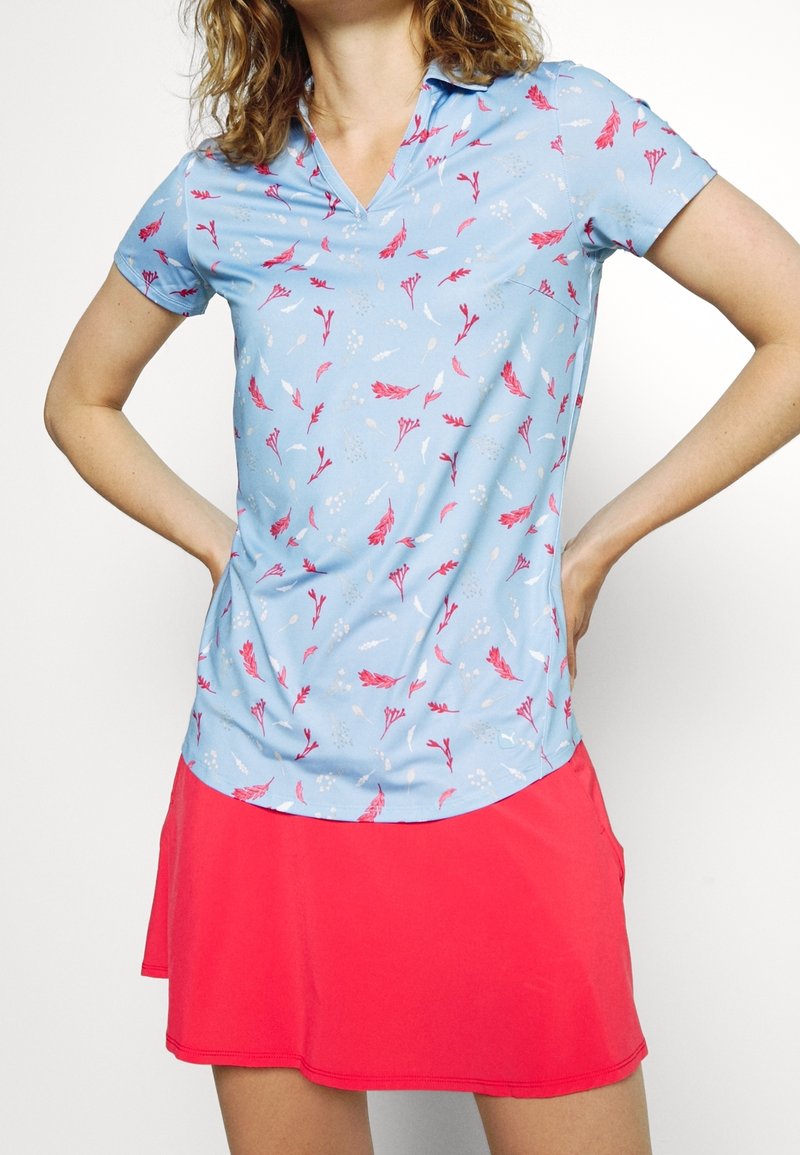 Femme portant une chemise bleu clair à col avec un imprimé floral rose et une jupe rouge vif, les mains posées sur les hanches.
