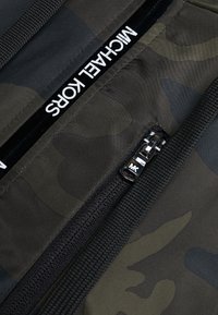 Close-up van een zwarte rits met het "MK" logo op een donker groen en zwart camouflage stof met "MICHAEL KORS" merknaam.