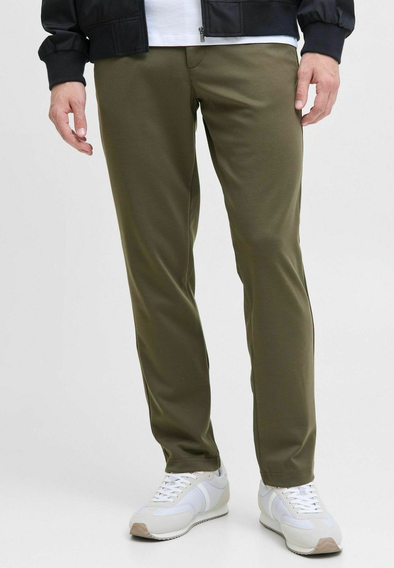 Pantalons slim en vert olive en tissu extensible, avec une texture lisse, associés à un t-shirt blanc uni et des baskets blanches.