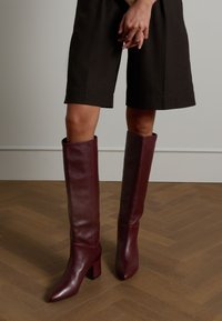 Bordeauxfarbene Leder-Stiefel bis ans Knie mit spitzem Zehenbereich, Blockabsatz und glatter Textur, getragen zu dunklen Shorts auf einem Holzboden.