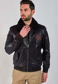 Homme ajustant le col d'une veste aviateur en cuir noir avec des patchs brodés, portant un pantalon cargo beige devant un fond uni.