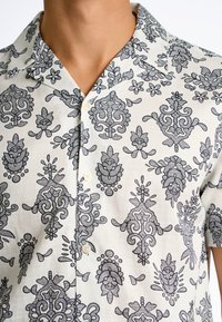 Camisa de manga corta hecha de tejido ligero, con un estampado floral y paisley negro sobre un fondo crema, cuello con puntas y botones.
