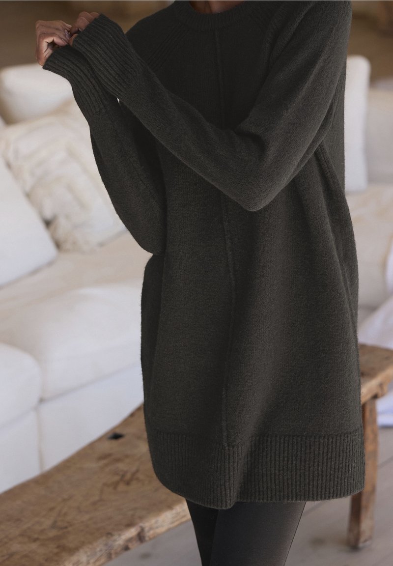 Personne portant un pull en maille surdimensionné de couleur sombre avec des poignets et un ourlet côtelés, ajustant la manche dans un salon confortable avec des canapés blancs et une table en bois.