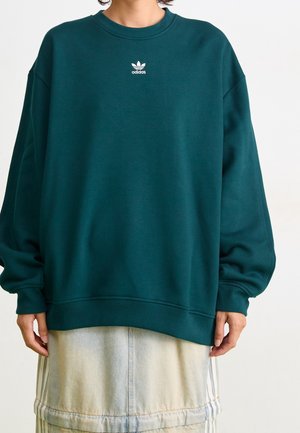 Persoon draagt een donkergroene Adidas-trui met wit logo, gecombineerd met een lichte denimrok met subtiele verticale strepen.