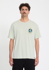 Mann mit Schnurrbart, der ein hellgrünes T-Shirt mit Globusgrafik und schwarze Hose trägt und vor einem schlichten weißen Hintergrund steht.