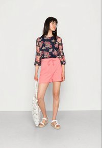 Blusa floral com mangas balão, fundo azul-marinho, combinada com calções coral claros. A modelo segura uma mala de tote branca com padrão, vestindo sandálias brancas.