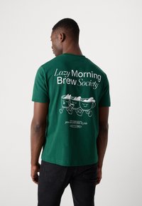 Grønn bomull T-skjorte med hvit tekst og tre illustrerte kaffekopper på baksiden, rund hals og korte ermer.