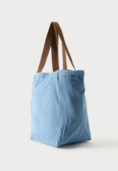 Sac cabas en tissu bleu clair avec des anses marron, debout sur un fond blanc.