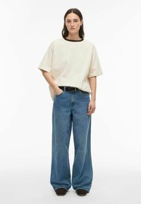 Ženska s ravnimi rjavimi lasmi nosi preveliko bež majico, ohlapne modre kavbojke s črnim pasom in temne čevlje, stoji pred belim ozadjem.