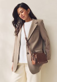 Blazer marron clair sur une chemise blanche, pantalon crème, et un petit sac bandoulière marron avec des fermetures éclair et des accents dorés. Textures lisses.