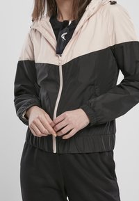 Giacca color block nera e rosa chiaro con zip, polsini elastici e cappuccio regolabile, realizzata in tessuto leggero e morbido.