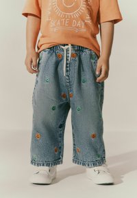 Denim bikses ar elastīgu jostu, ar daudzkrāsainiem smaidošu seju iz�šuvumiem, brīvs piegriezums un apģērbti ar baltām slip-on kedām.