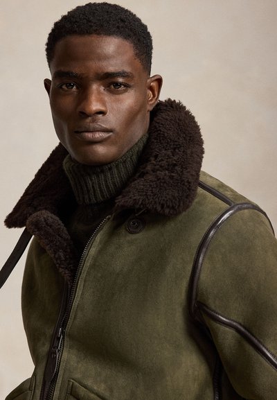 Mann trägt dunkelgrüne Jacke mit großem braunem Fellkragen und dunkelgrünem geripptem Rollkragenpullover, schaut geradeaus vor neutralem Hintergrund.
