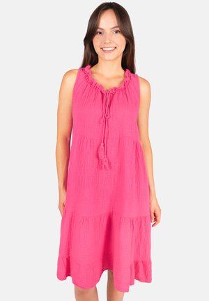 Jeune femme souriante portant une robe sans manches rose vif avec un col volanté et des liens à pompons.