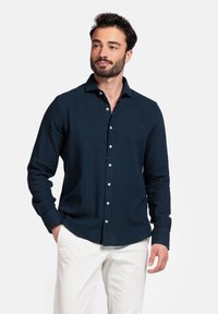 Marineblauw overhemd met lange mouwen en knopen, met een kraaghals en textuurstof. Voorzien van witte knopen en een rechte zoom. Wordt gedragen met witte broeken.