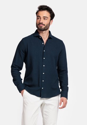 Marineblauw overhemd met lange mouwen en knopen, met een kraaghals en textuurstof. Voorzien van witte knopen en een rechte zoom. Wordt gedragen met witte broeken.