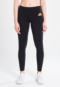 Svart tights med en glatt tekstur, høy midje og en fargerik logoakseent på hoften. Sammen med hvite treningssko.