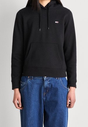 Person, die einen schwarzen Hoodie mit Vordertasche und kleinem Logo trägt, kombiniert mit lockeren blauen Jeans, vor einem schlichten hellen Hintergrund stehend.