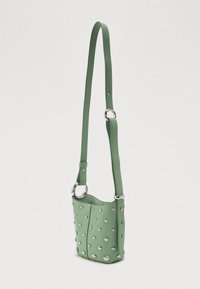 Πράσινη δερμάτινη τσάντα τύπου μπucket με ασημί καρφιά και ρυθμιζόμενο μακρύ λουρί, που παρουσιάζεται σε ένα απλό λευκό φόντο.