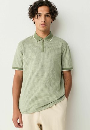 Camisa polo verde con patrón texturizado, de manga corta y cuello con media cremallera. Presenta acentos en blanco contrastantes en el cuello y las mangas.