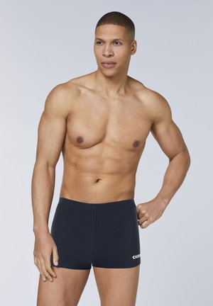 Fit man with short hair posing in black swim trunks against a plain light background, looking slightly to the side.