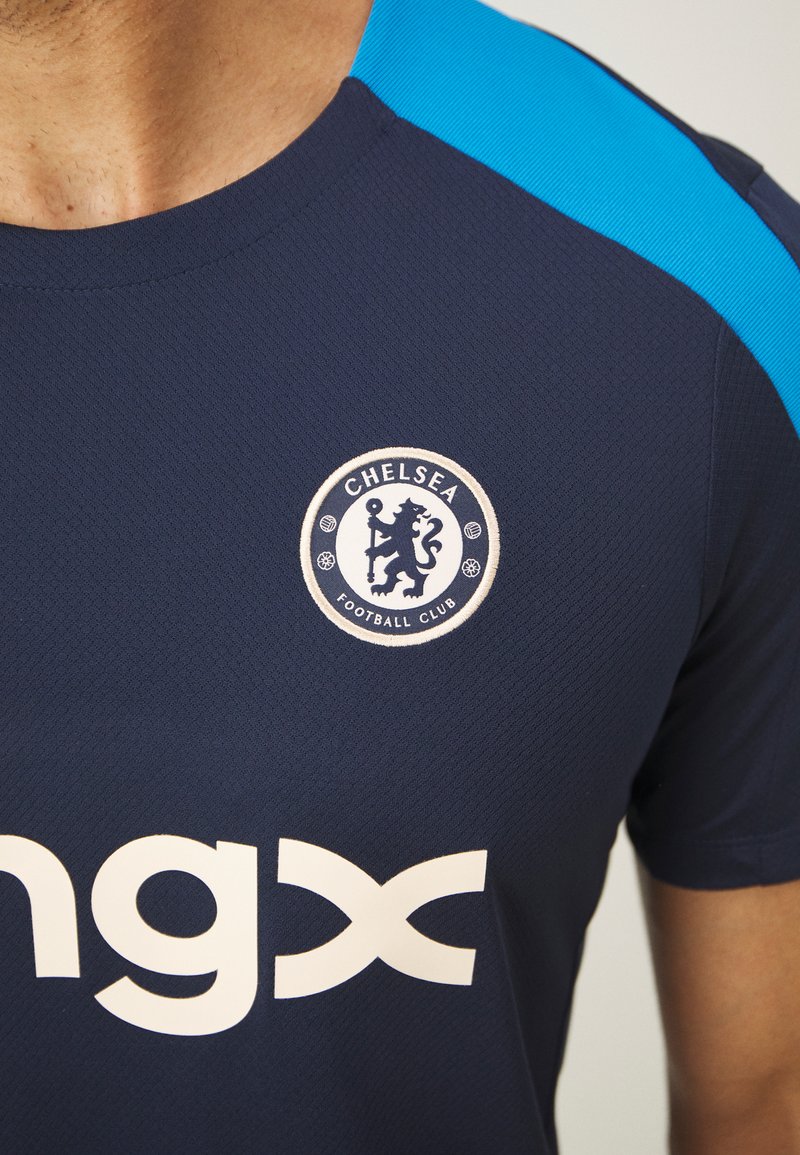 Granatowa koszulka piłkarska z jasnoniebieskimi akcentami na ramionach, z okrągłym logo Chelsea FC i dużym białym napisem "ngx".