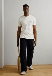 Tricou crem cu mânecă scurtă și un logo pe piept, asortat cu pantaloni negri largi și adidași gri, purtând o geantă de tote bej.