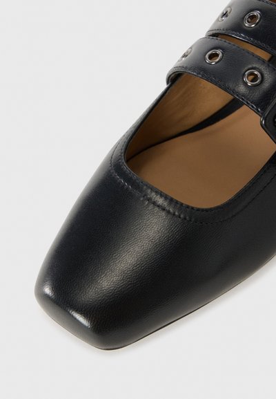Chaussure en cuir noir avec un bout carré, dotée de deux larges bretelles avec des œillets en métal circulaires. Texture lisse avec une légère brillance.