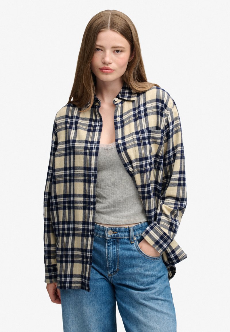 Jeune femme aux longs cheveux bruns porte une chemise à carreaux beige et marine sur un débardeur gris et un jean bleu, une main dans la poche.