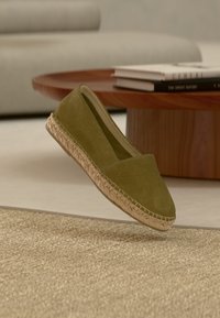 Une espadrille vert olive avec une semelle en jute cousue flottant au-dessus d'un tapis beige, avec une table en bois et des livres en arrière-plan.