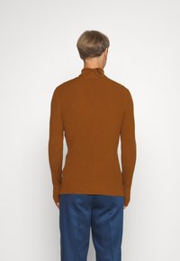 Maglione a collo alto marrone a coste con colletto asimmetrico; il modello indossa pantaloni di raso blu. Tessuto strutturato, design aderente.