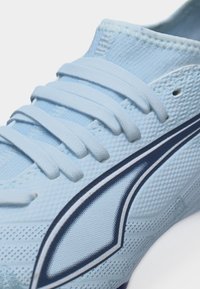 Približen posnetek svetlo modrega športnega čevlja z teksturirano tkanino, usklajenimi vezalkami in temno modrim ter belim ukrivljenim črtastim vzorcem ob strani.