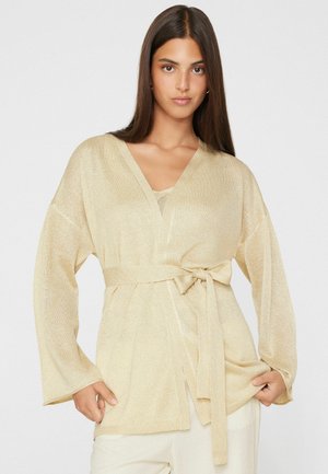 Donna con lunghi capelli castani che indossa un cardigan beige lavorato a maglia, legato in vita, e pantaloni color crema, in piedi davanti a uno sfondo semplice.