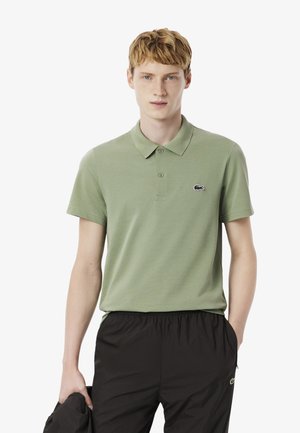 Joven vestido con una camiseta polo verde claro y pantalones negros, sosteniendo una chaqueta negra en una mano contra un fondo blanco.