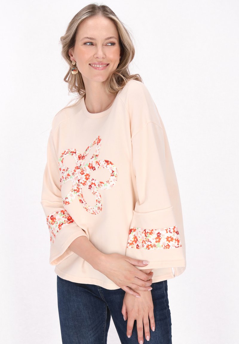 Pfirsichfarbener, übergroßer Sweatshirt mit floralem Muster an den Ärmeln und der Vorderseite, runder Ausschnitt und weiche Stofftextur.